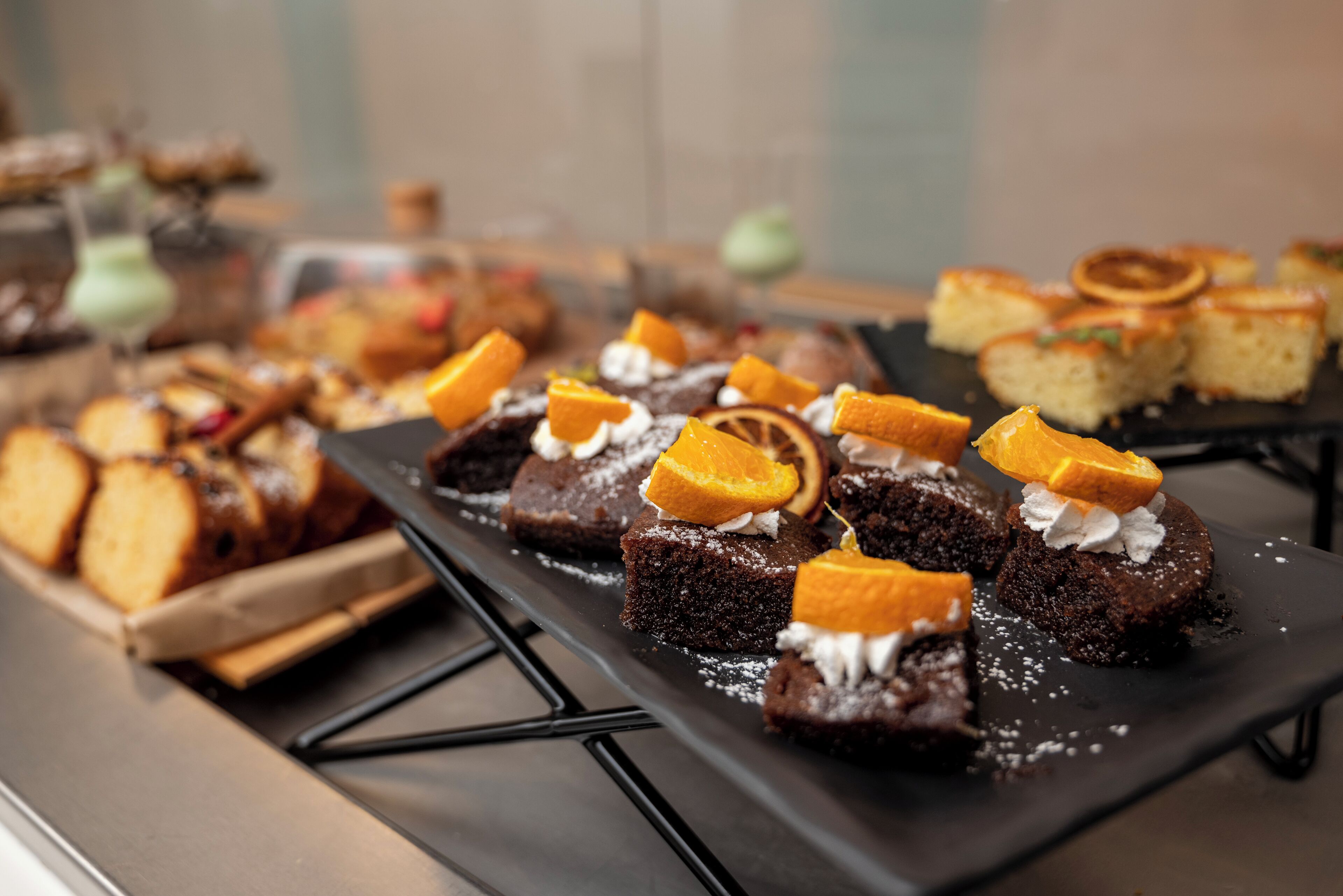 Une variété de desserts présentés sur un plateau, notamment des brownies et des gâteaux, dont certains sont décorés de tranches d’orange et de crème.