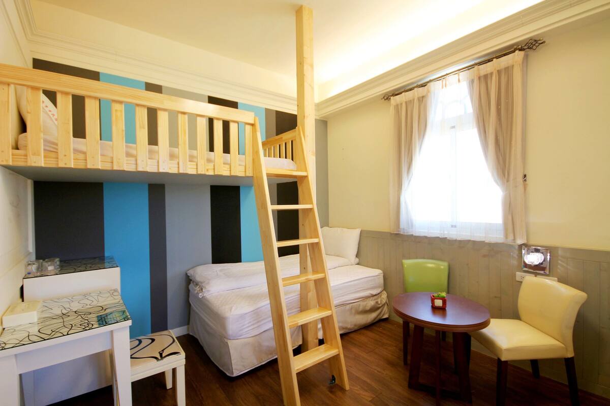 A room with bunk beds, a wooden ladder, a small table, a chair, and a window with curtains.