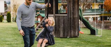 A man and a child on a swing in a park with a playground and a building in the background.