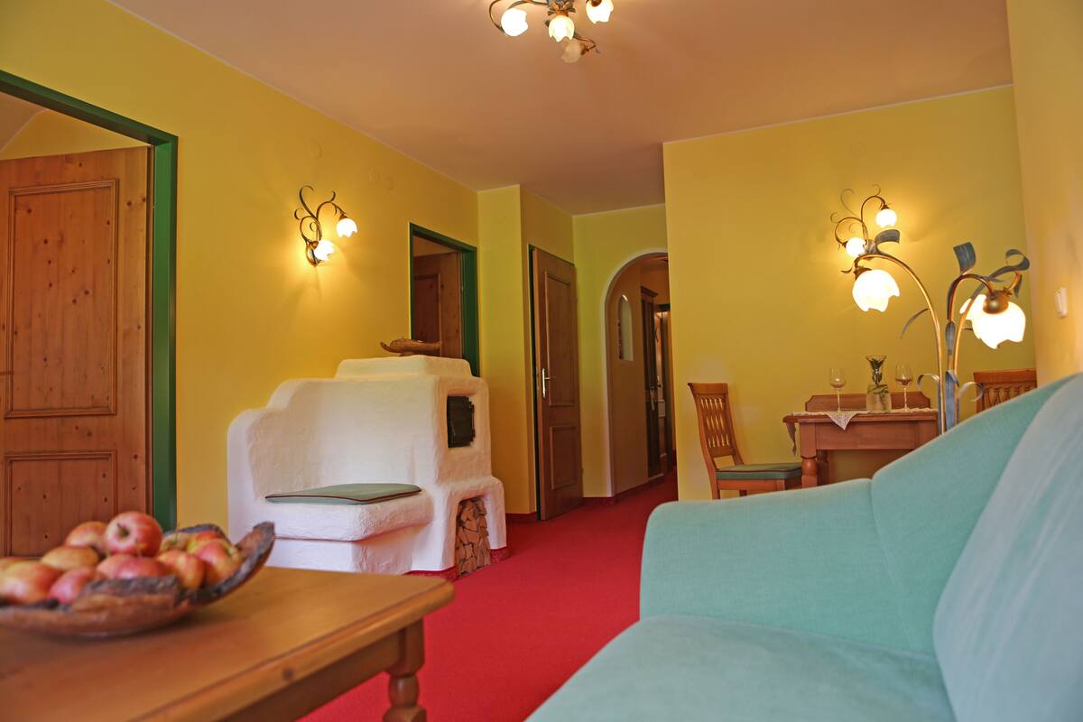 A room with a wooden door, a white armchair, a teal sofa, a wooden table with a fruit bowl, and wall-mounted light fixtures.