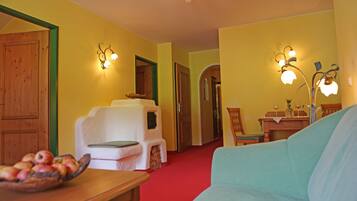 A room with a wooden door, a white armchair, a teal sofa, a wooden table with a fruit bowl, and wall-mounted light fixtures.