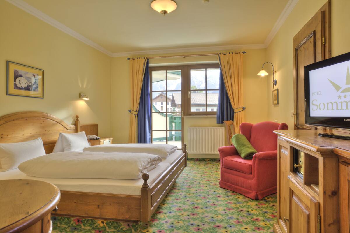 A hotel room with two beds, a television, a red armchair, and a window with curtains.
