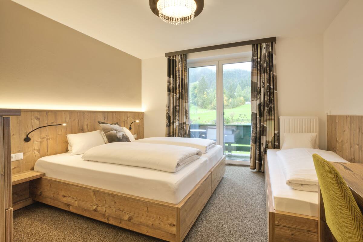 A bedroom with a large bed, a smaller bed, a desk, and a view of a green landscape through the window.