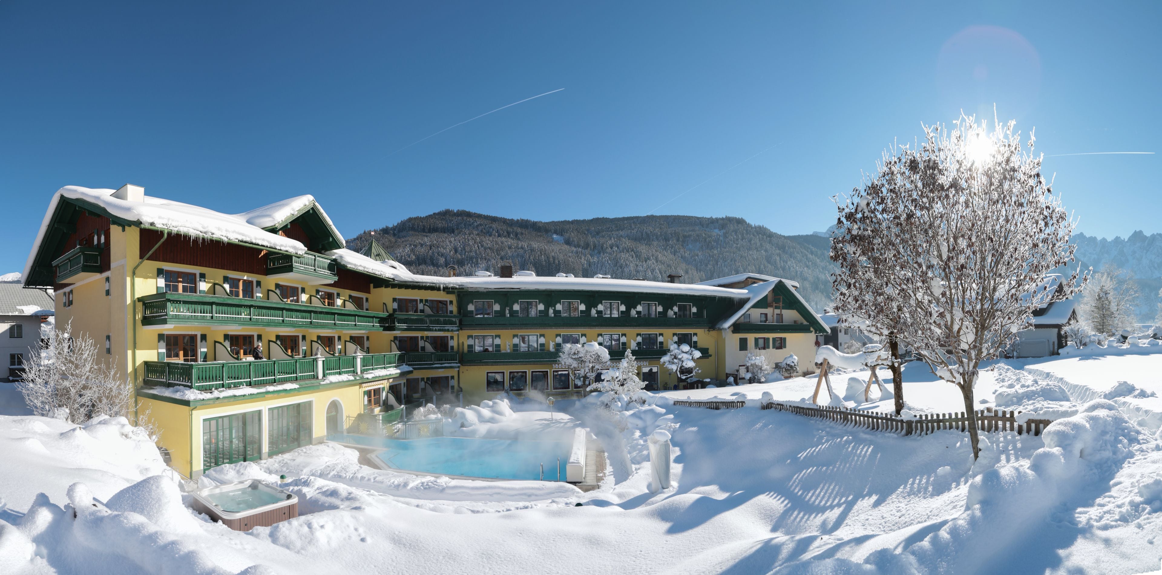A hotel with a pool area surrounded by snow, a mountain backdrop, and a clear sky.