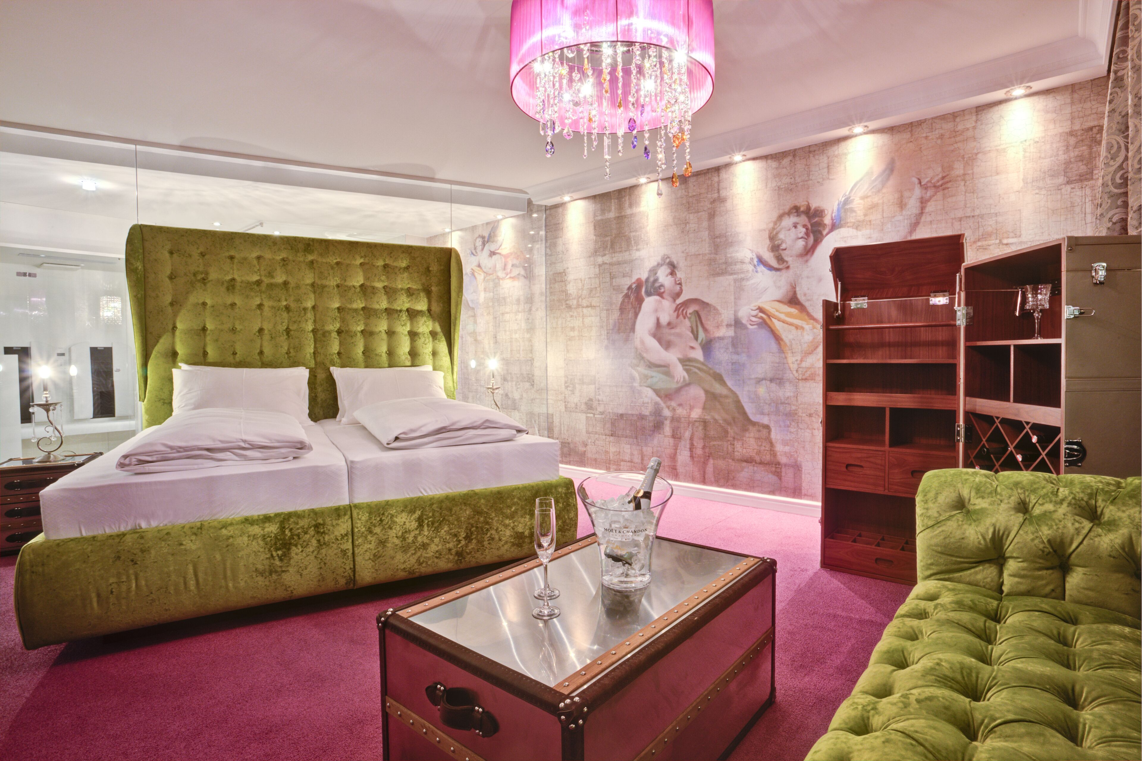 A bedroom with a green upholstered headboard, a glass coffee table, a wooden cabinet, and a pink chandelier.