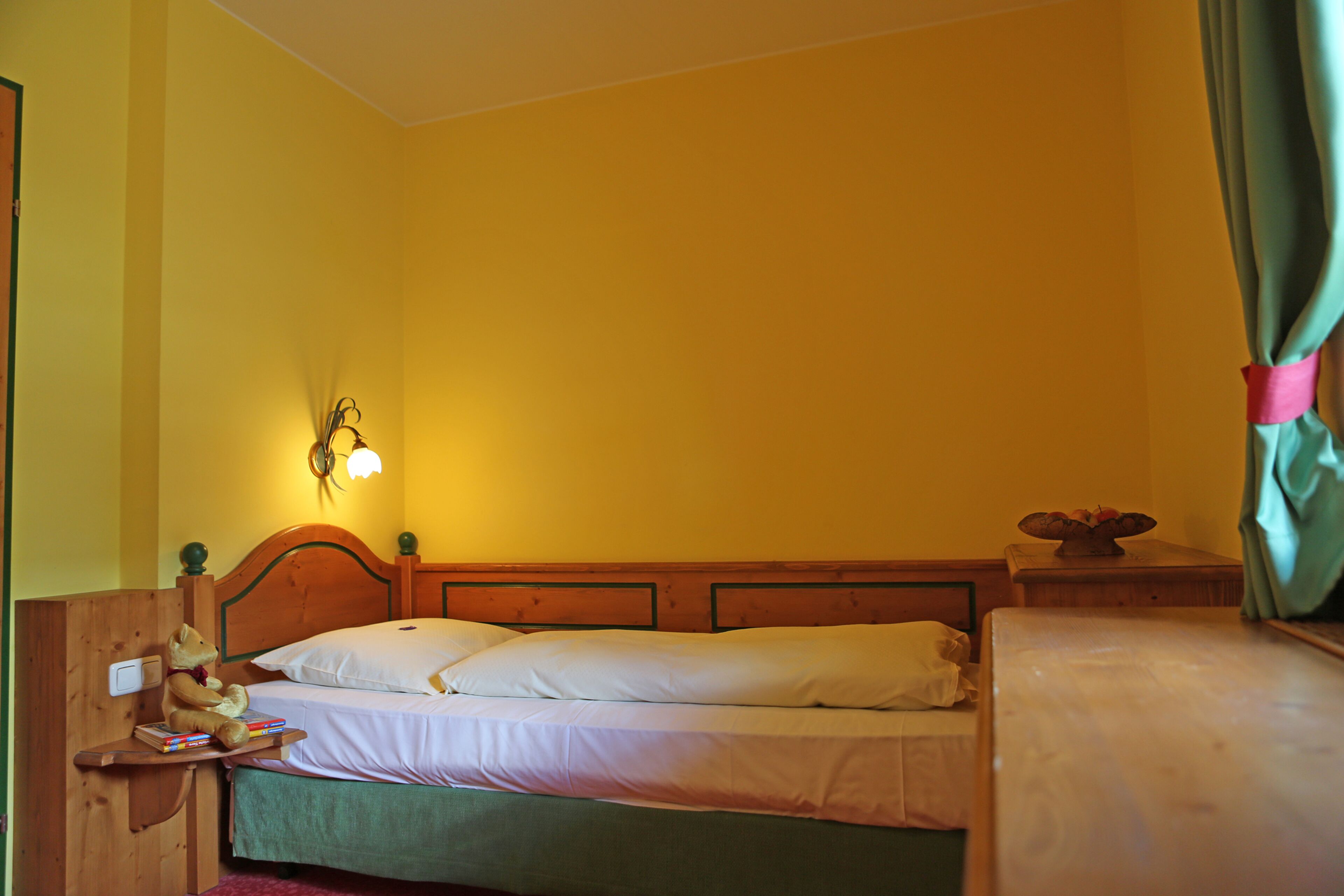 A single bed with a wooden headboard, a bedside table with a lamp, and a teddy bear on the nightstand.