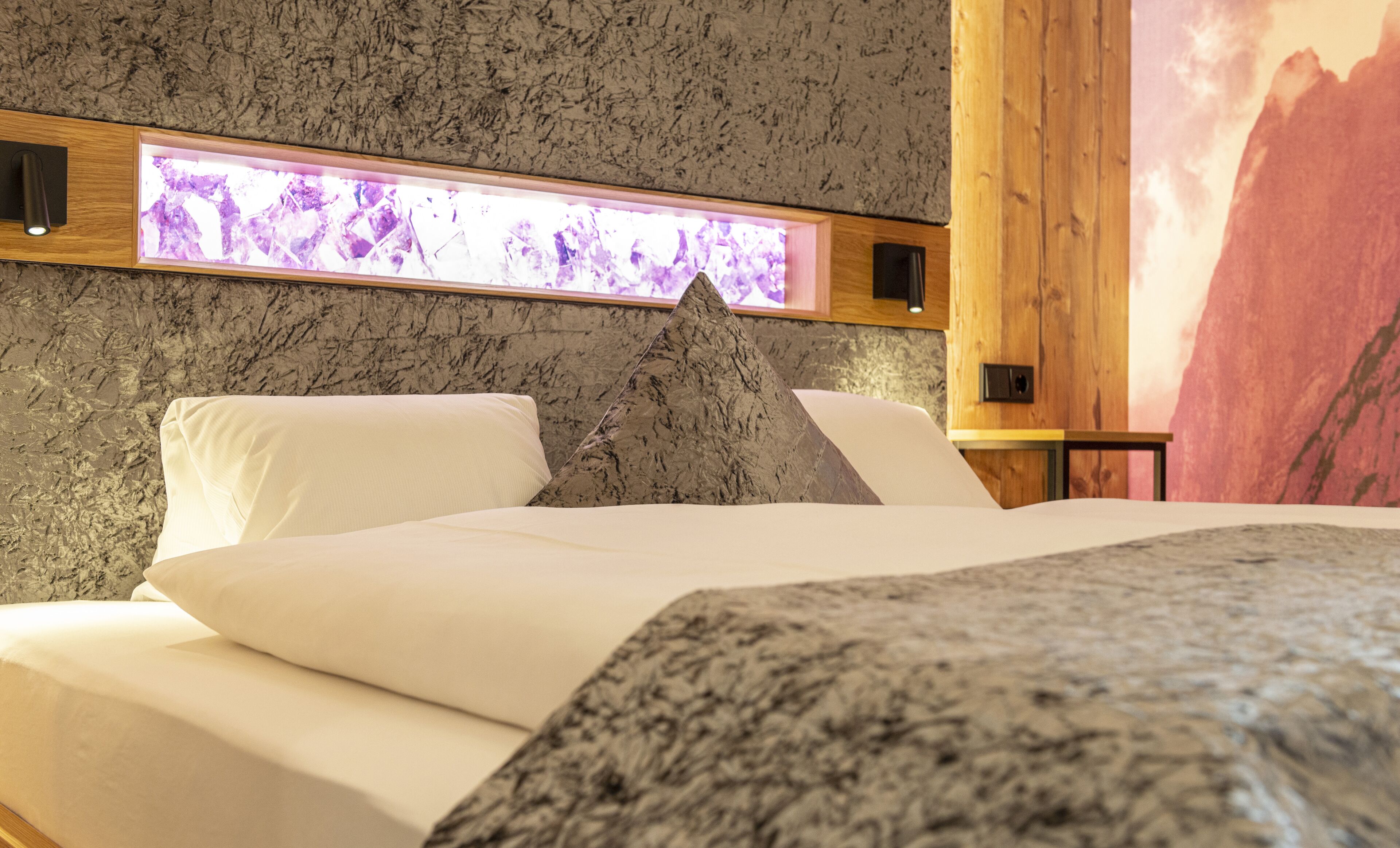 A bed with white linens and a gray headboard in a room with a textured wall and a wooden panel.