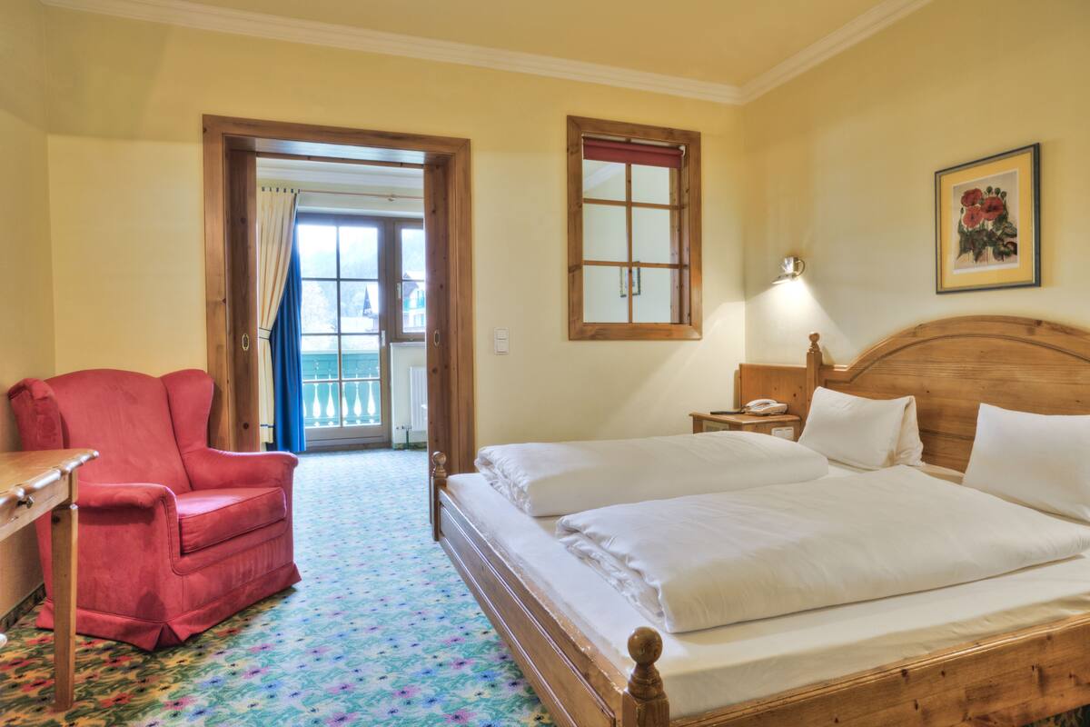 A hotel room with a wooden bed, a red armchair, a small table, and a window with a view.