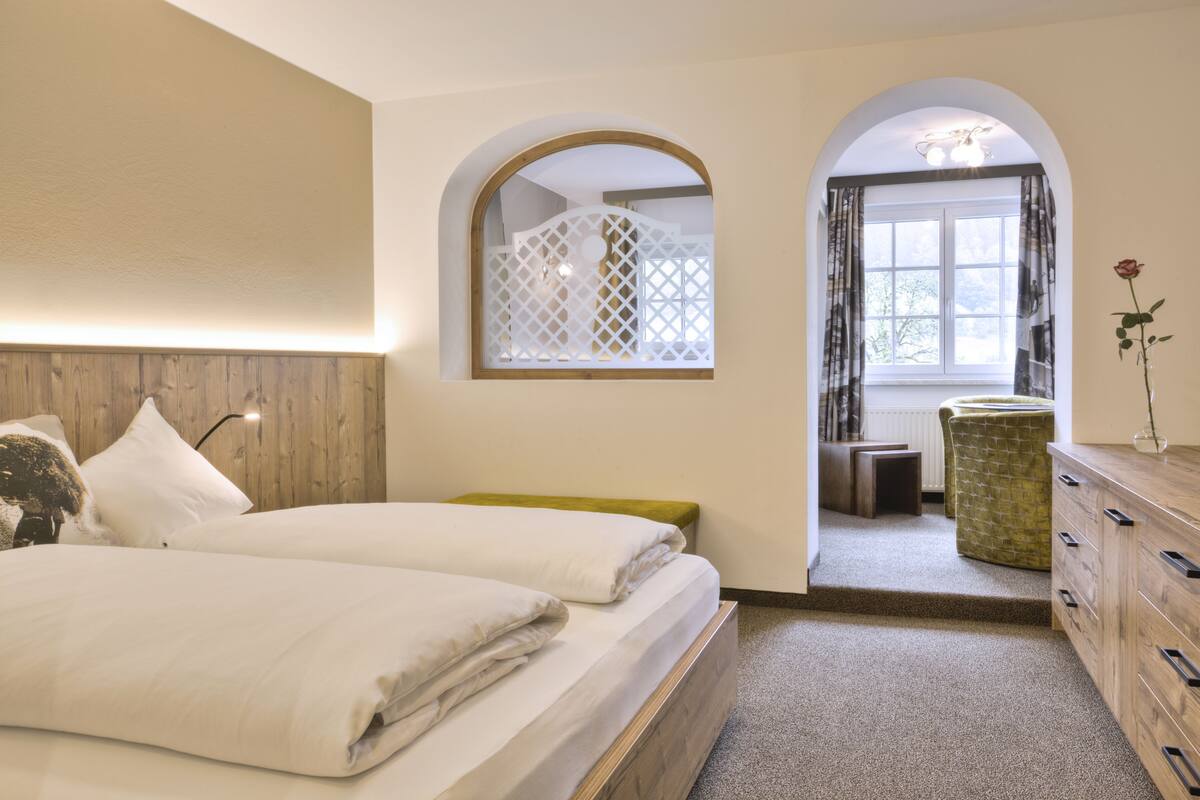A bedroom with a wooden headboard, a bed with white bedding, a green bench, a wooden dresser, and a window with curtains.