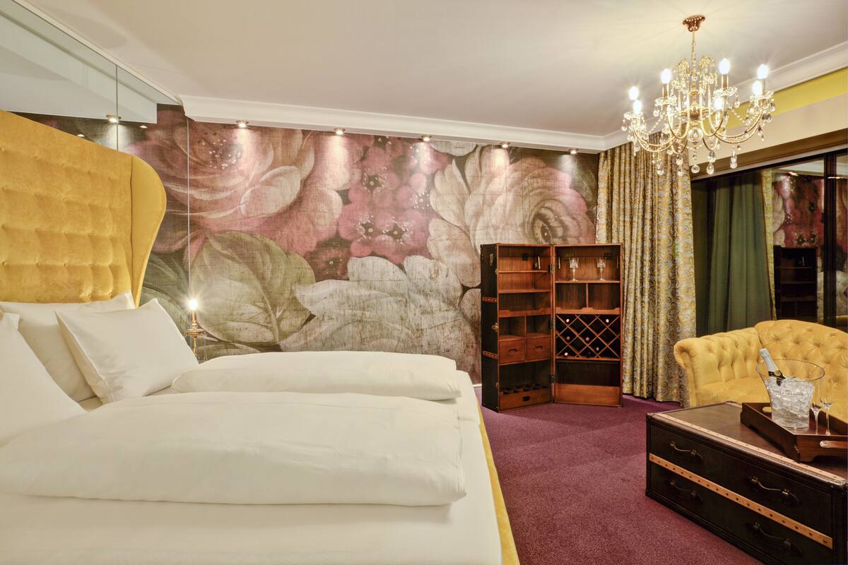 A bedroom with a large bed, a wooden bookshelf, a yellow armchair, and a chandelier.