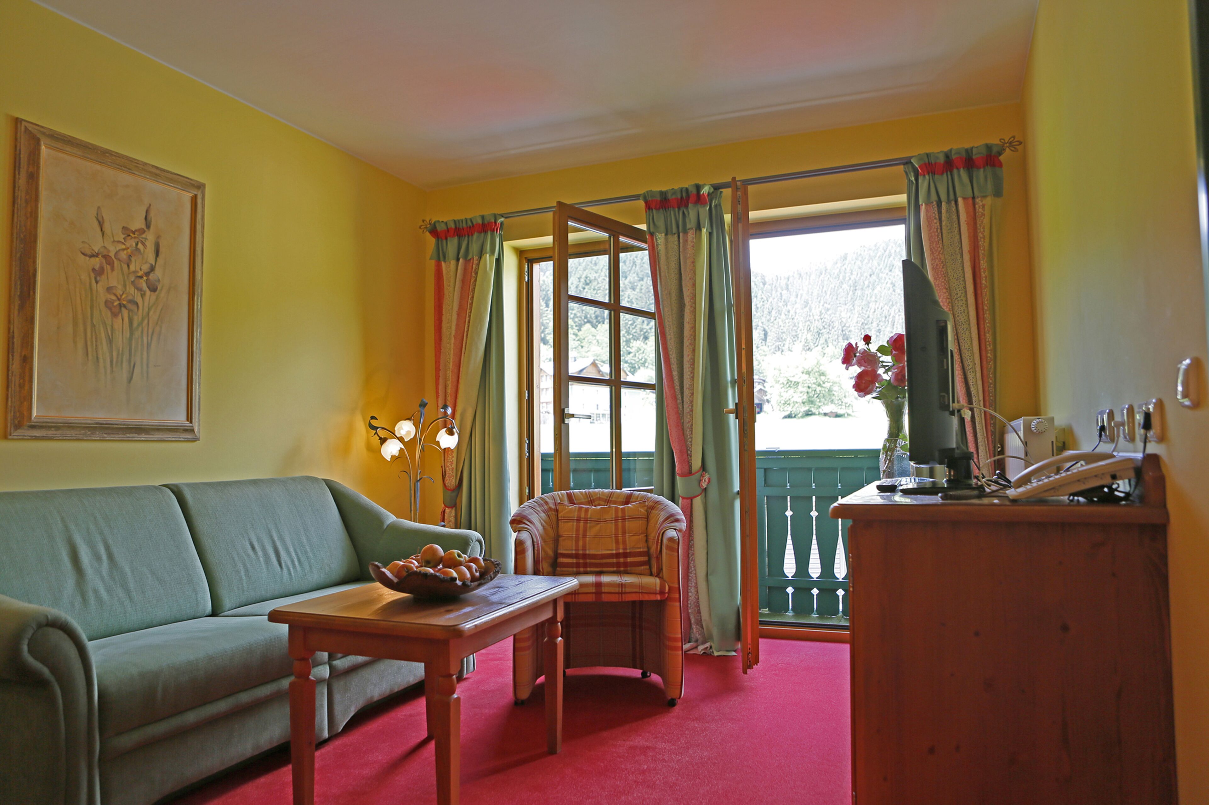 A room with a sofa, a table with a fruit bowl, a plaid armchair, a wooden desk with a telephone, and a window with curtains.