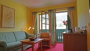 A room with a sofa, a table with a fruit bowl, a plaid armchair, a wooden desk with a telephone, and a window with curtains.
