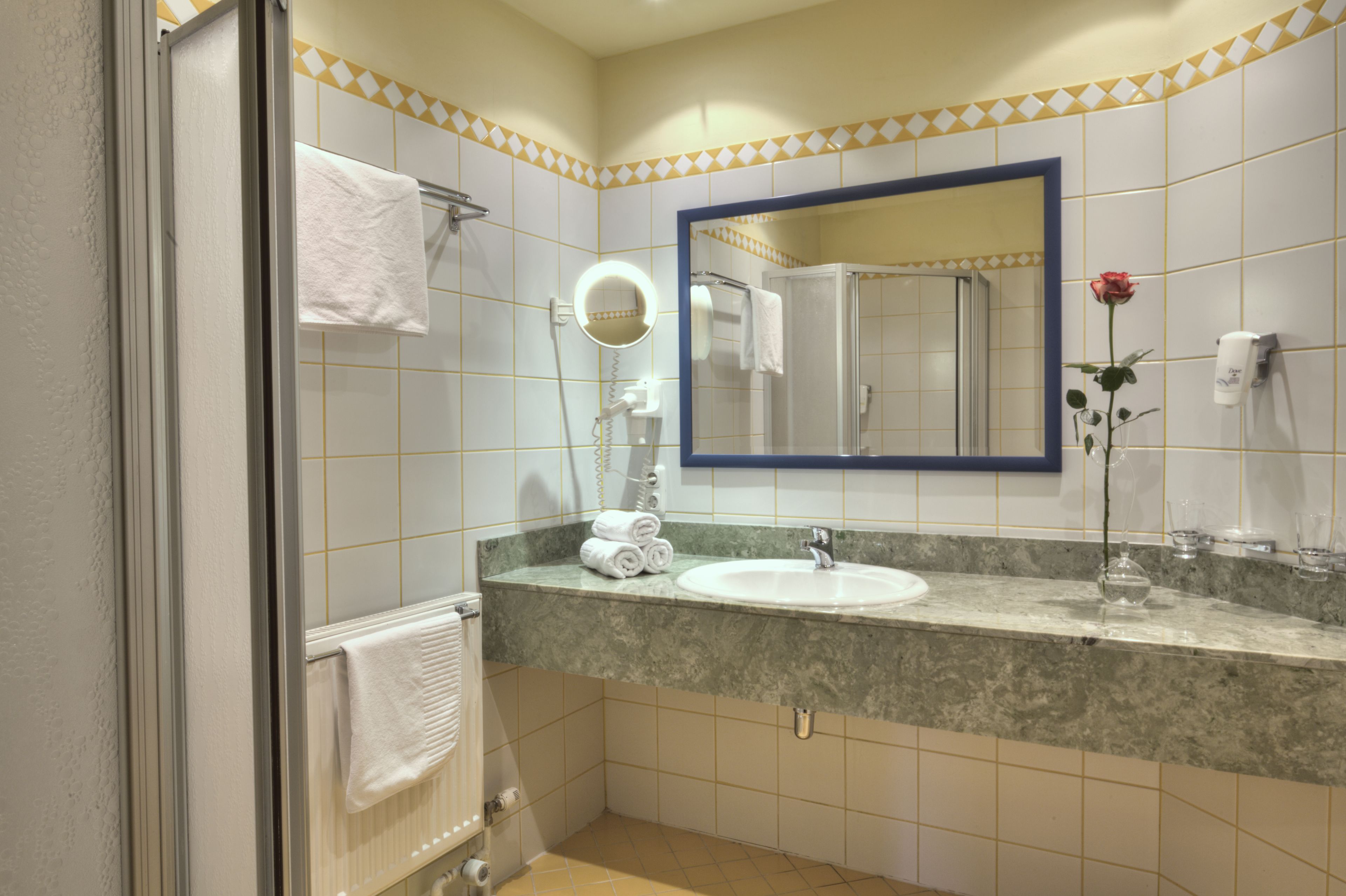 Un baño con lavamanos, espejo, toallero y una rosa en un florero.