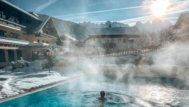 A person in a clear blue pool with steam rising, surrounded by buildings and snow-covered trees. - Hotel Sommerhof (Gosau)
