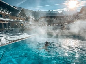 A person in a clear blue pool with steam rising, surrounded by buildings and snow-covered trees. - Hotel Sommerhof (Gosau)