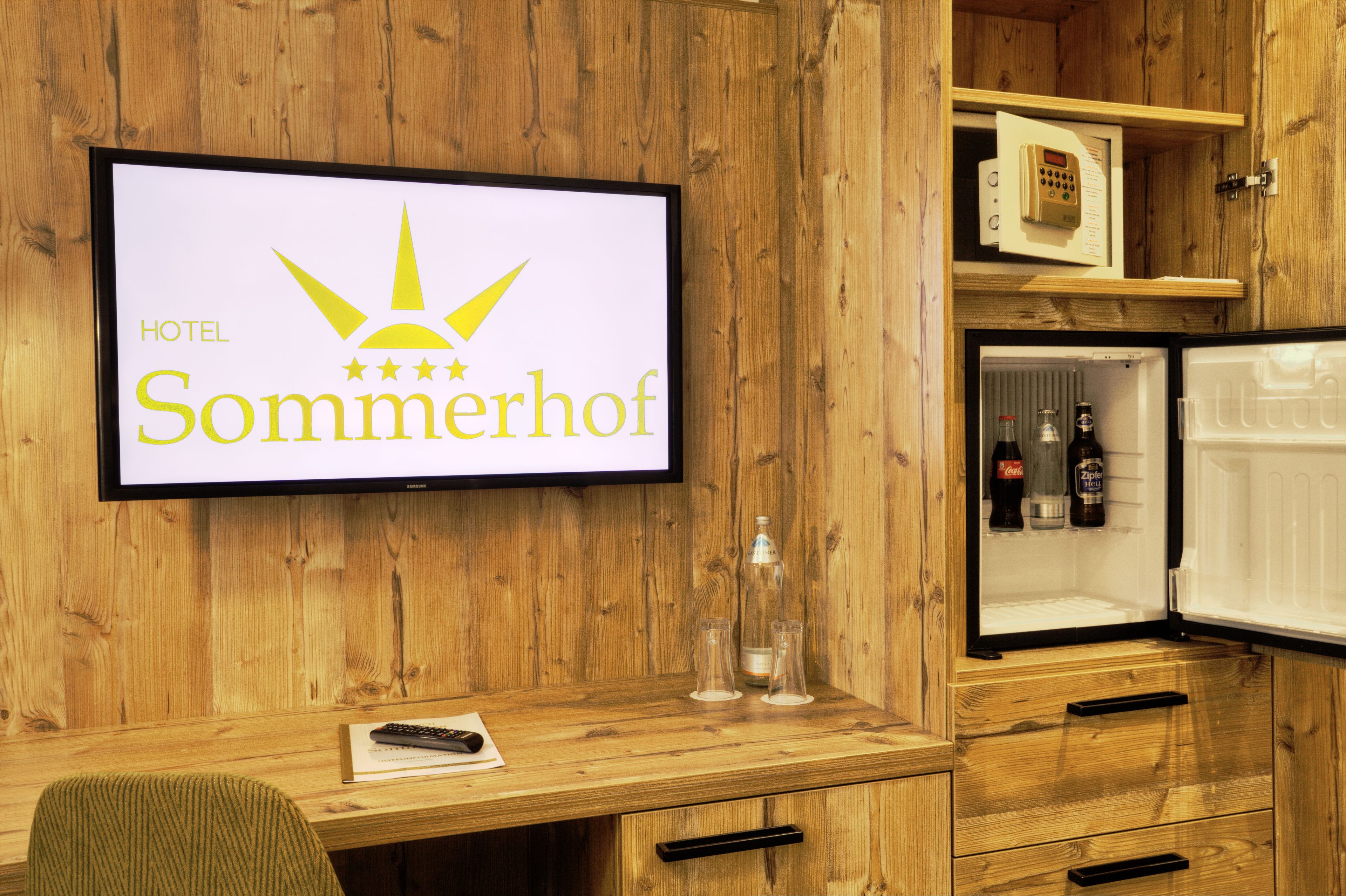 A hotel room with a wooden interior, a television displaying the hotel's name, a minibar, and a desk with a remote control.