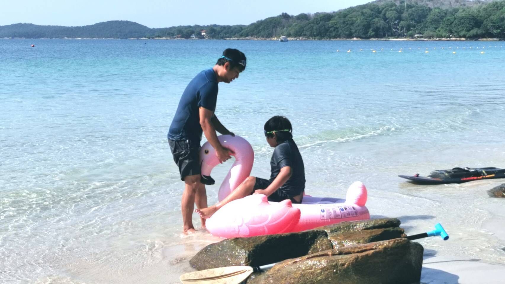 Escena de playa con agua cristalina, una persona de pie sobre una roca y otra sentada en un flotador rosa con forma de flamenco.