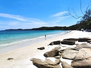 Une plage avec de gros rochers au premier plan, un ciel bleu dégagé et une personne qui marche sur le sable.