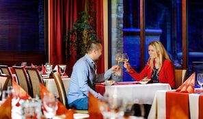Ein Paar erhebt Weingläser in einem Restaurant mit roten Vorhängen und einer Pflanze im Hintergrund.