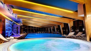 Uma piscina interior moderna com água azul cristalina, vigas de madeira no teto e espreguiçadeiras ao redor.