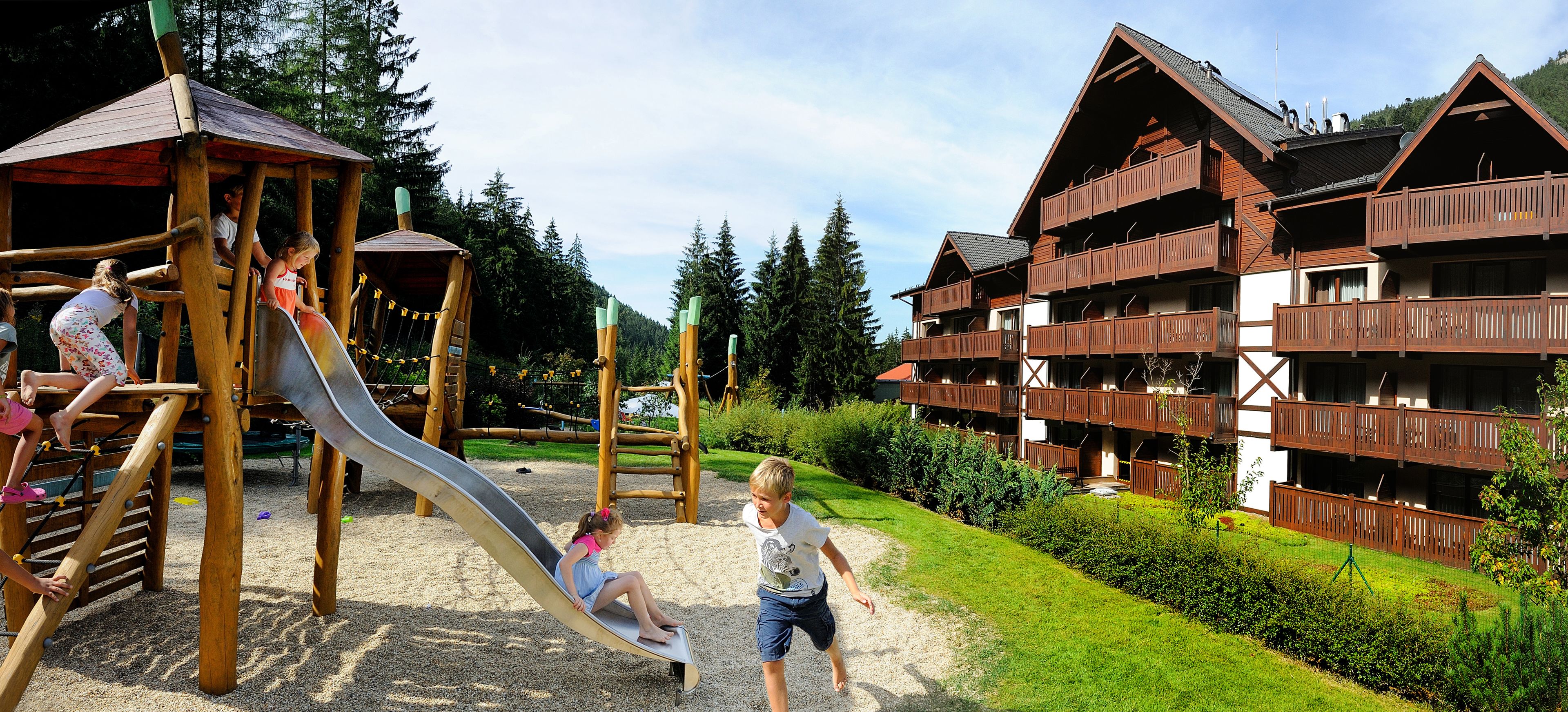 A playground with a slide and climbing structures, surrounded by a well-maintained garden and a chalet-style building with balconies.