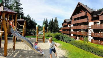 A playground with a slide and climbing structures, surrounded by a well-maintained garden and a chalet-style building with balconies.