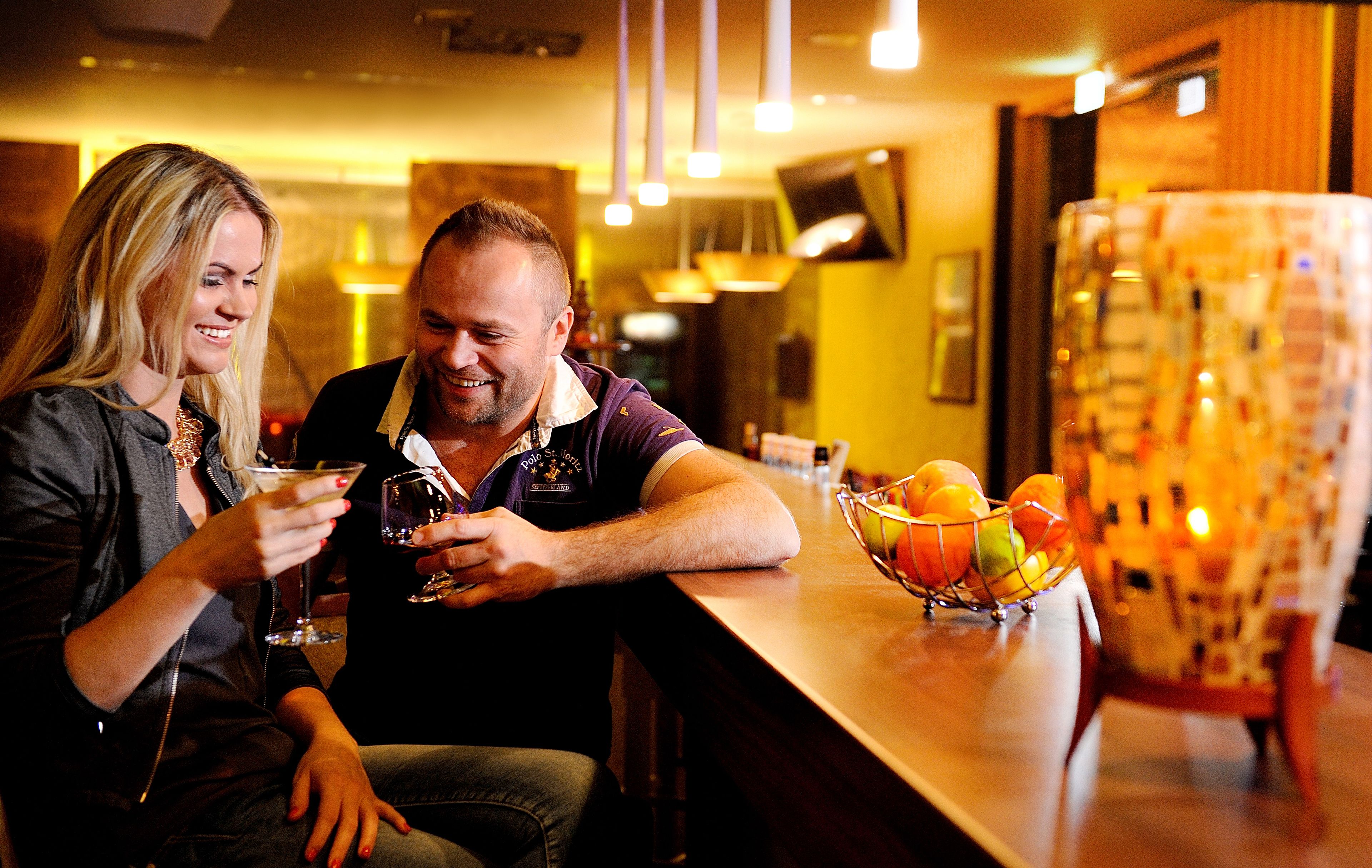 Una pareja sentada en un bar, con un frutero y botellas al fondo.