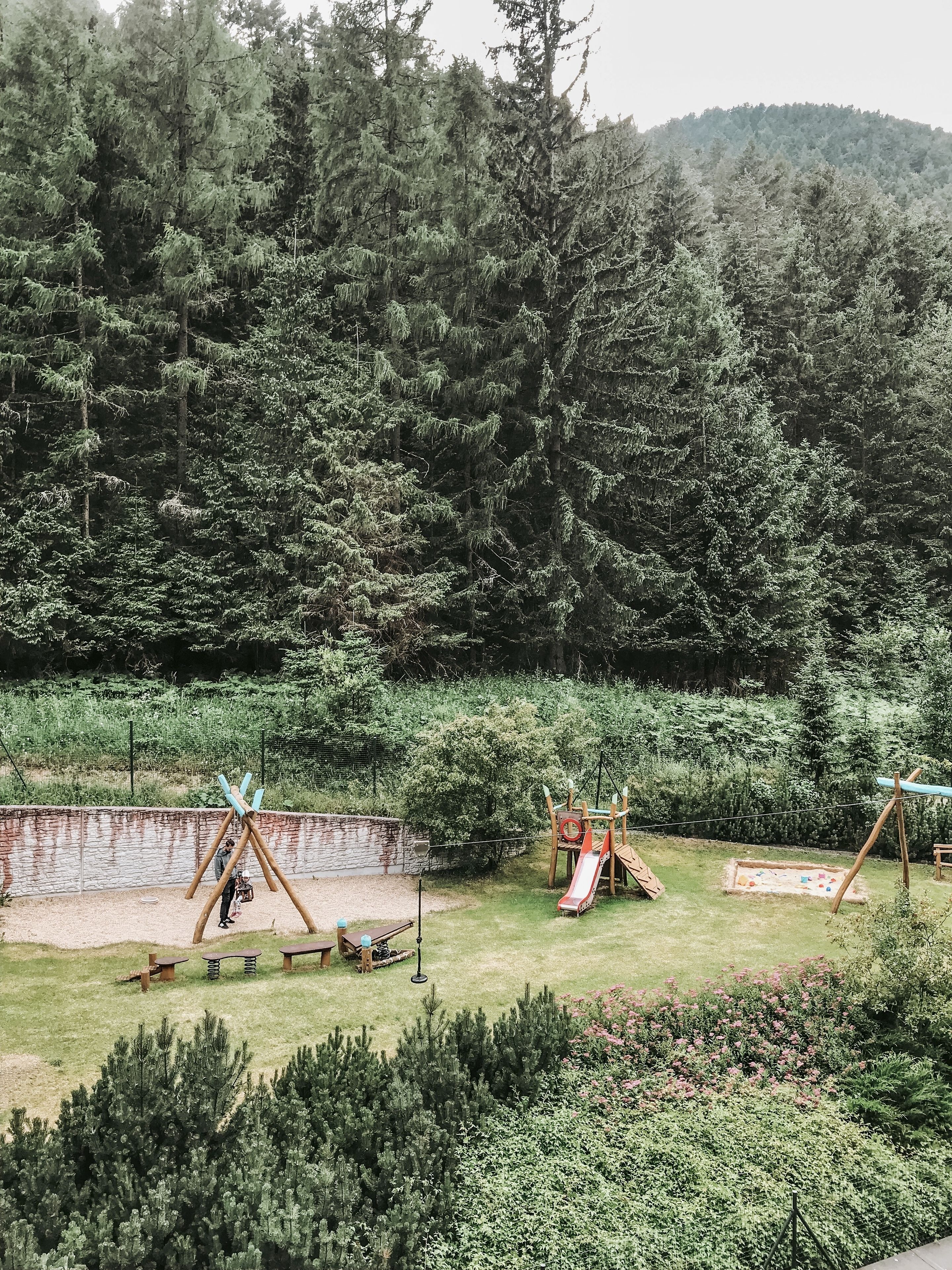 Un parque infantil con columpios y toboganes, rodeado de árboles frondosos y un muro de piedra.