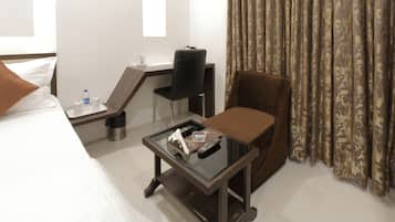 A hotel room with a bed, a desk with a chair, a small table with a remote control, and a window with curtains.