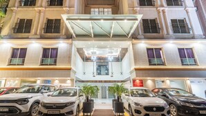 Entrada de um hotel com vários andares, com toldo de vidro, carros estacionados e plantas em vasos.