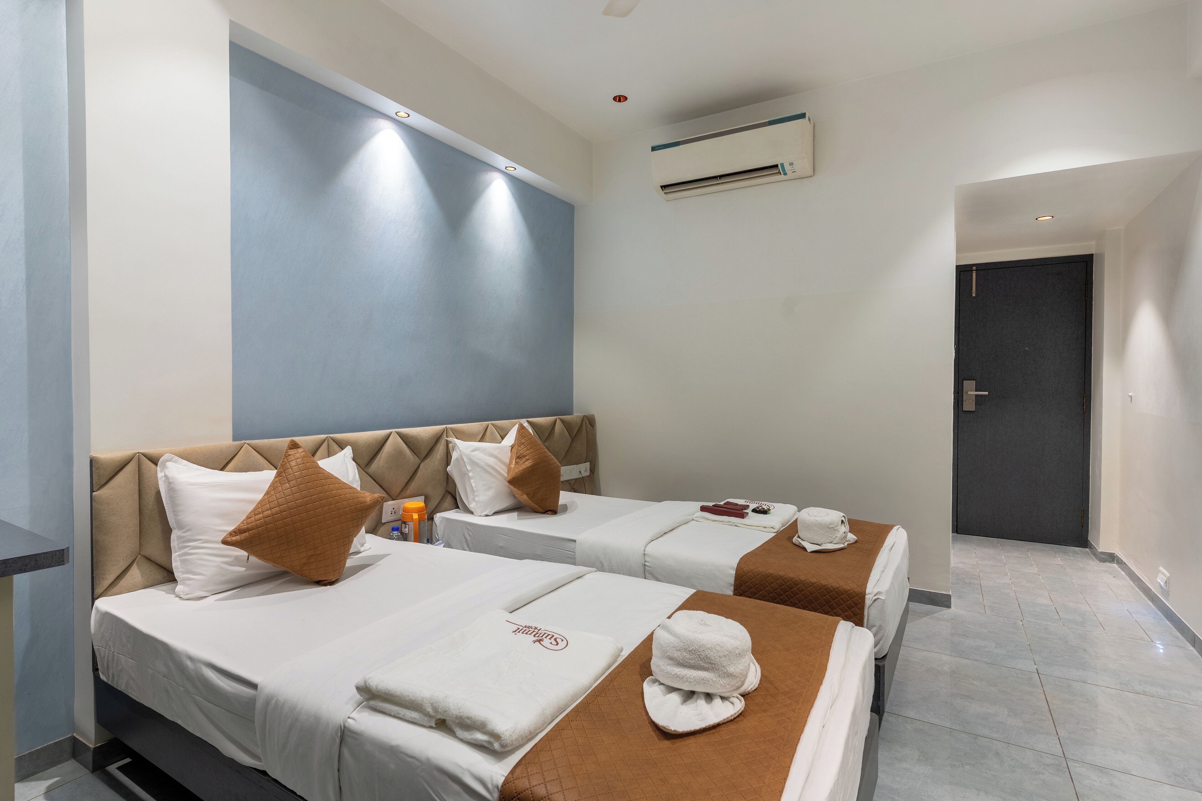 A hotel room with two beds, each with a brown headboard, white bedding, and a towel with a logo. There is a small table with a bottle and a remote control.