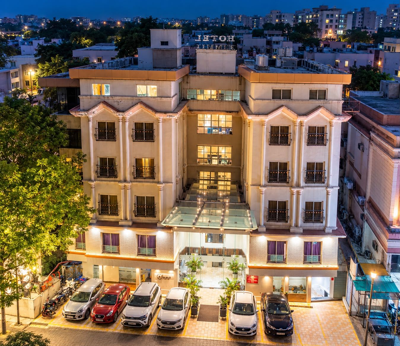 Hotel de varios pisos con un atrio central, un estacionamiento con varios automóviles y vista del paisaje urbano al anochecer.