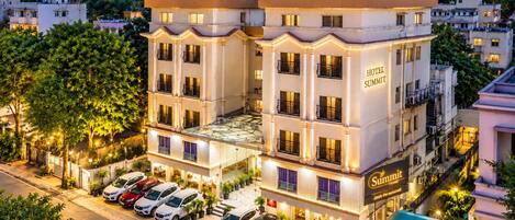 Ein mehrstöckiges Hotel mit markantem Eingang, einer Dachterrasse und einem Parkplatz mit mehreren Autos.