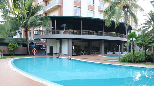 A swimming pool with a curved edge, surrounded by a tiled walkway. There are palm trees and a building with a balcony in the background.