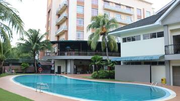 Una piscina con una silla de salvavidas y una tabla de salto, rodeada de palmeras y un edificio de varios pisos con balcones.