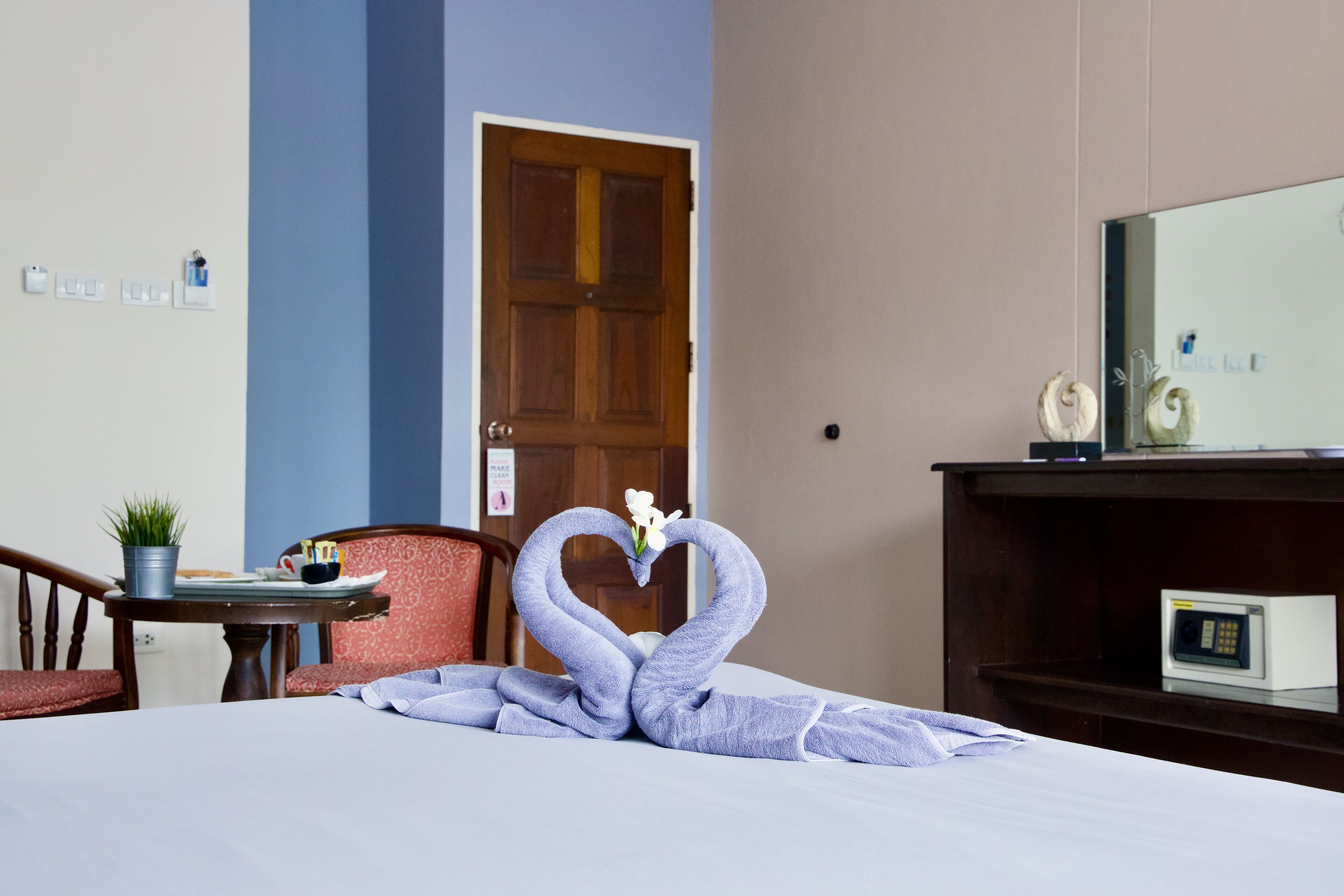 A hotel room with a bed, a desk with a computer, a chair, a door, and a wall-mounted shelf with decorative items.