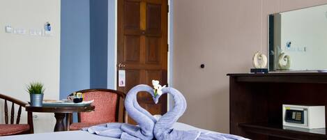 A hotel room with a bed, a desk with a computer, a chair, a door, and a wall-mounted shelf with decorative items.