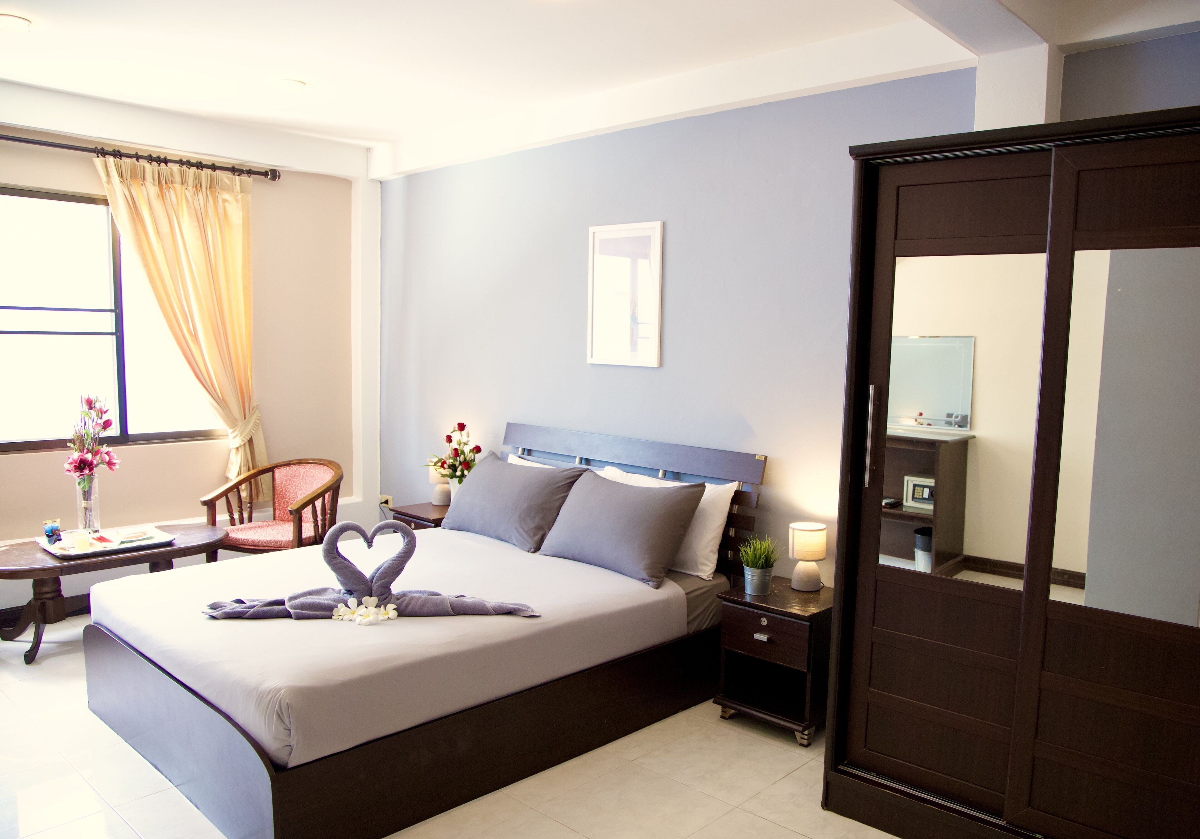 A hotel room with a large bed, a bedside table with a lamp, a chair, and a desk with a vase of flowers.