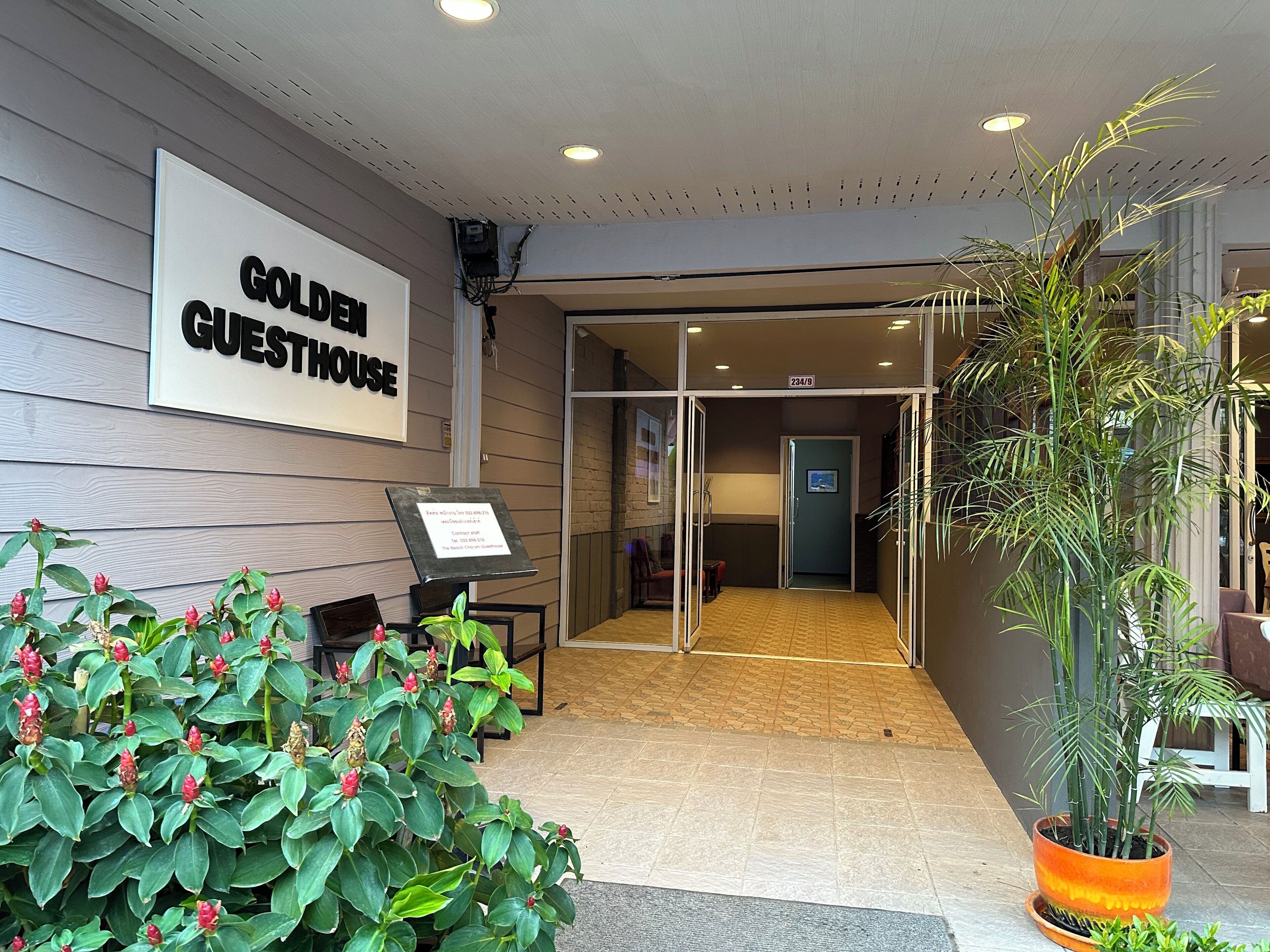 L’entrée d’un hôtel, avec une enseigne indiquant « GOLDEN GUESTHOUSE », un comptoir d’accueil, une plante en pot et un sol carrelé.