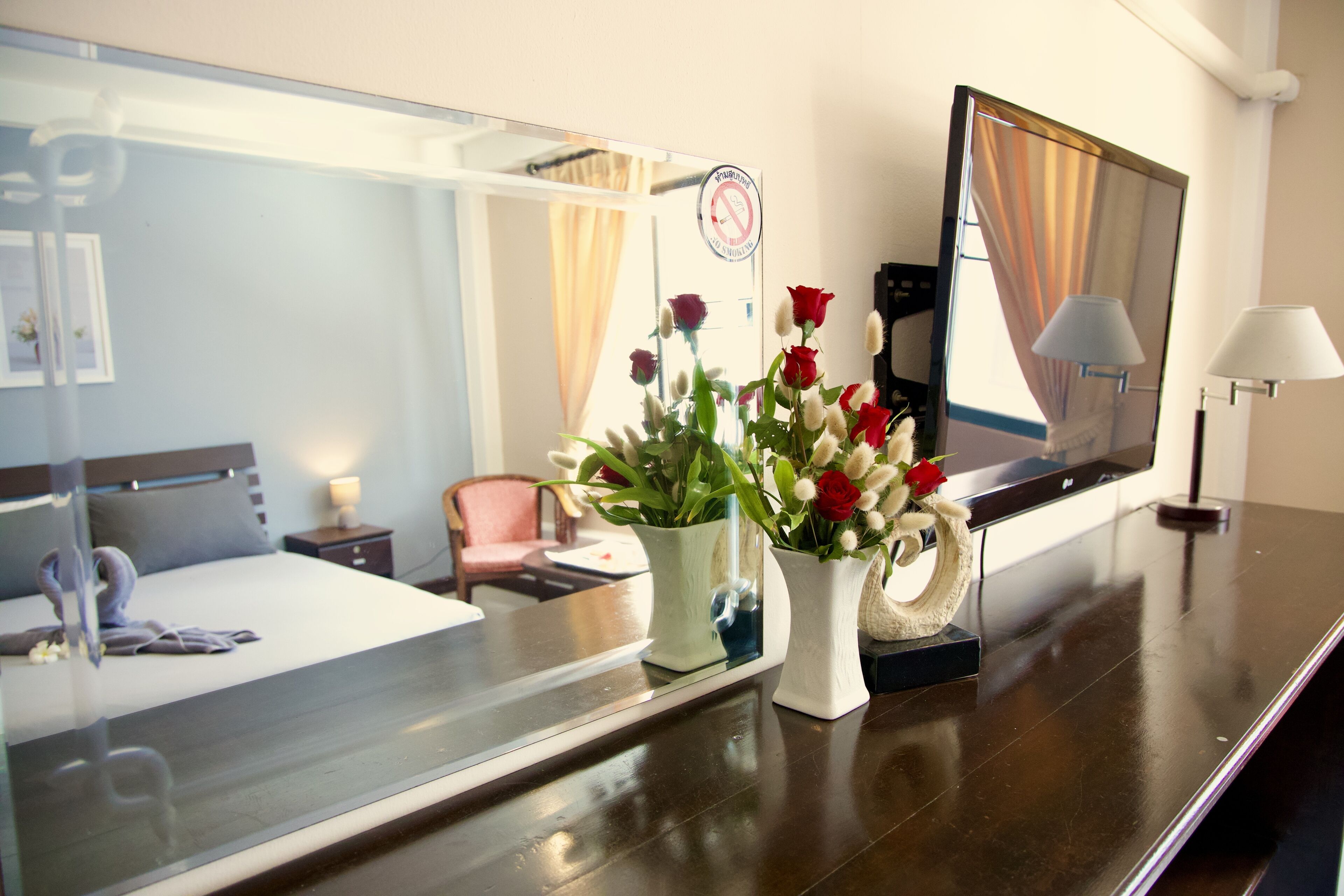 A hotel room with a bed, a bedside table with a lamp, a mirror, and a vase with flowers.