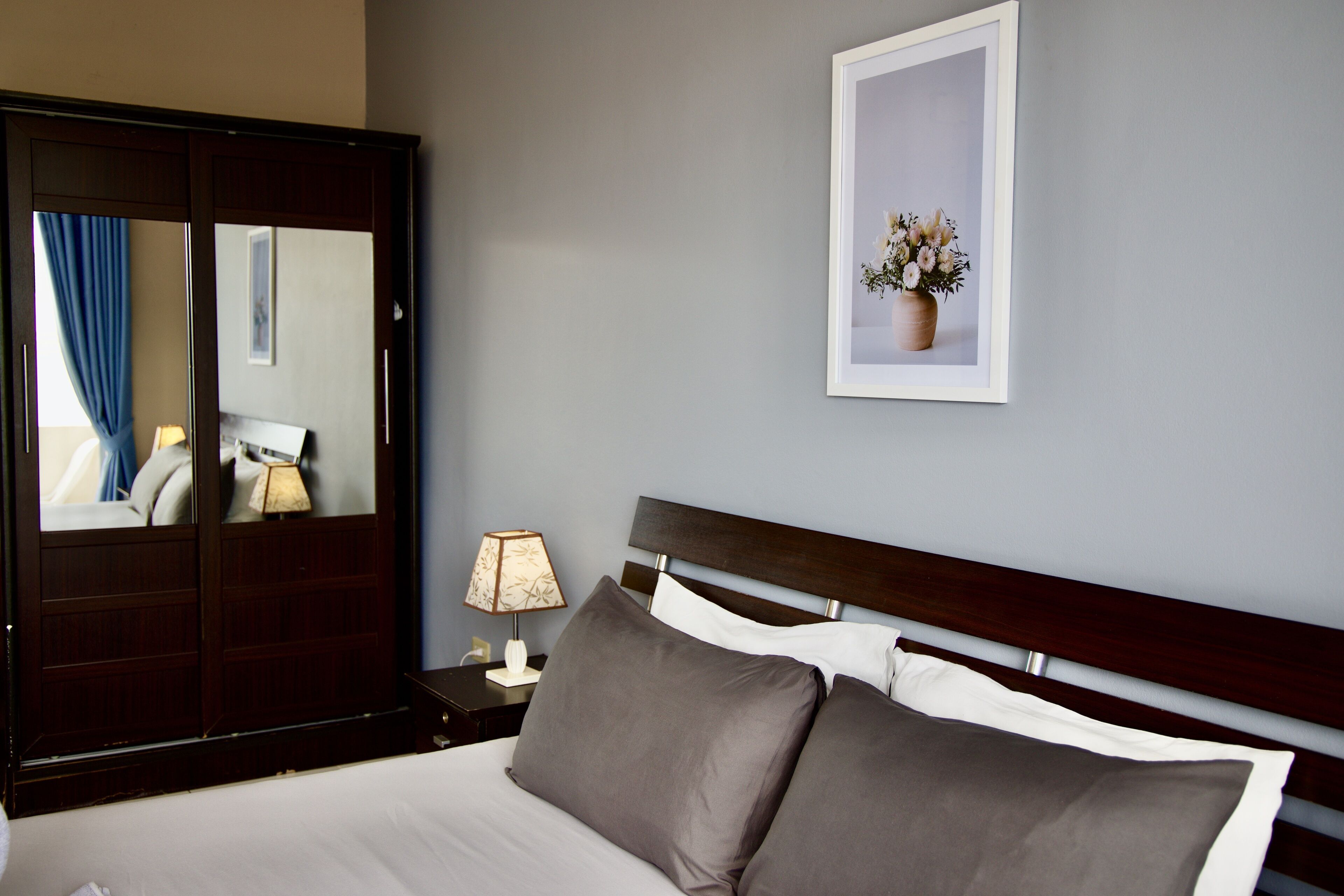 A hotel room with a large bed, a nightstand with a lamp, a mirror, a framed picture on the wall, and a wardrobe.