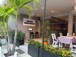 Un área de estar al aire libre con mesas y sillas, un letrero que dice 'GOLDEN GUESTHOUSE' y varias plantas en macetas.