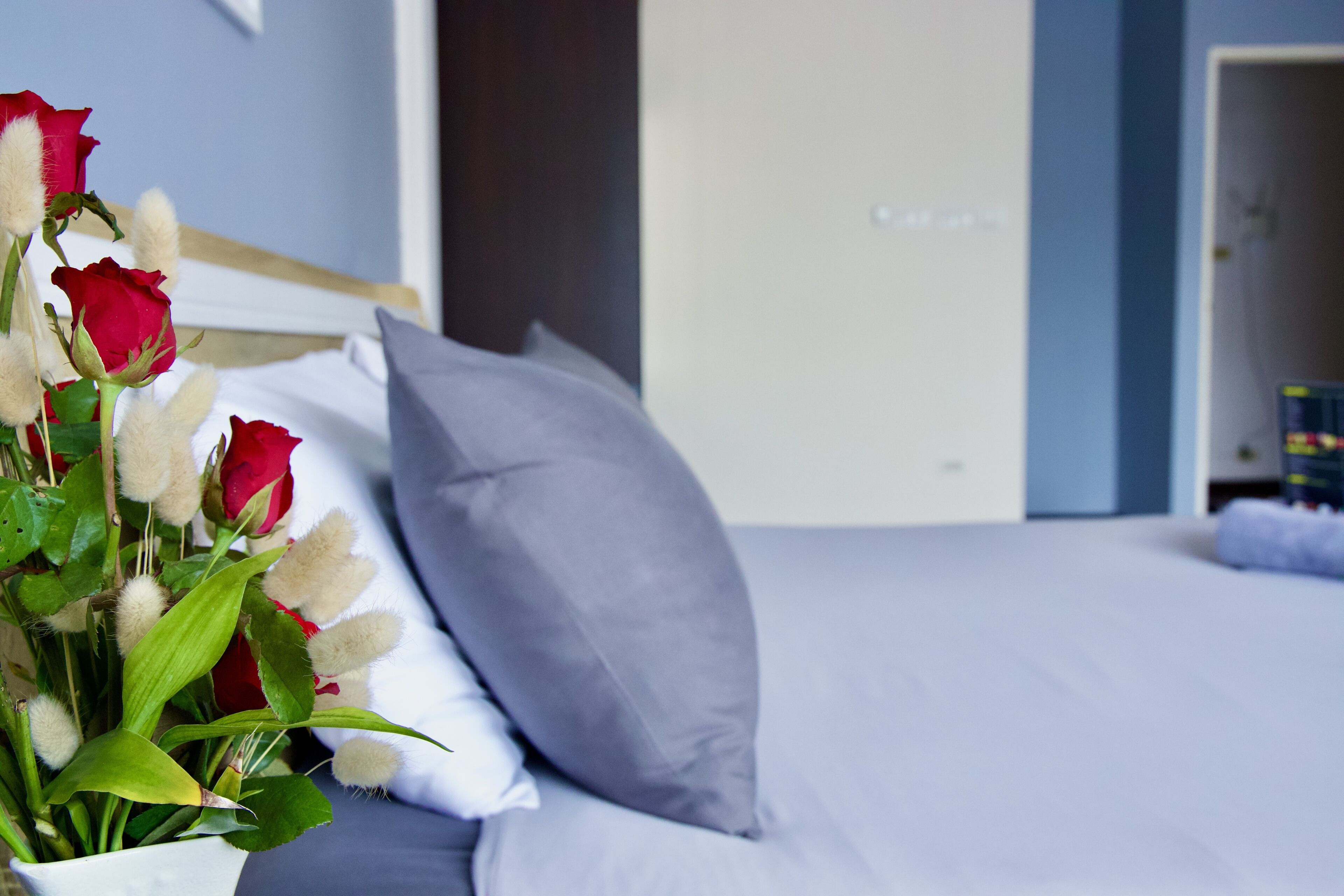 Un lit avec un oreiller gris et un bouquet de roses rouges dans un vase blanc.