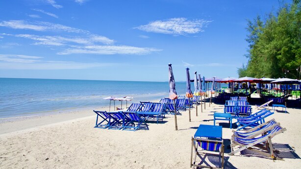 Een strand met rijen blauw-witte gestreepte ligstoelen en parasols, een zandstrand en een kalme zee onder een heldere lucht.