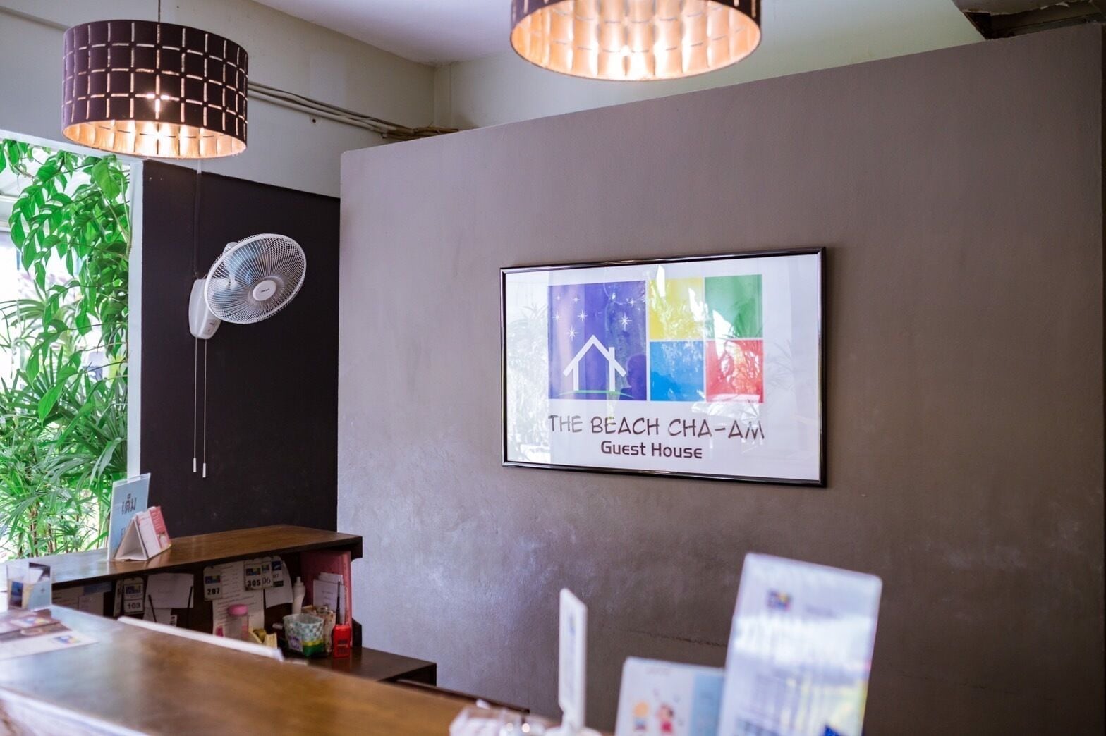 Zona de recepción con un cartel enmarcado que dice "The Beach Cha-Am Guest House", un ventilador y un mostrador con diversos objetos.