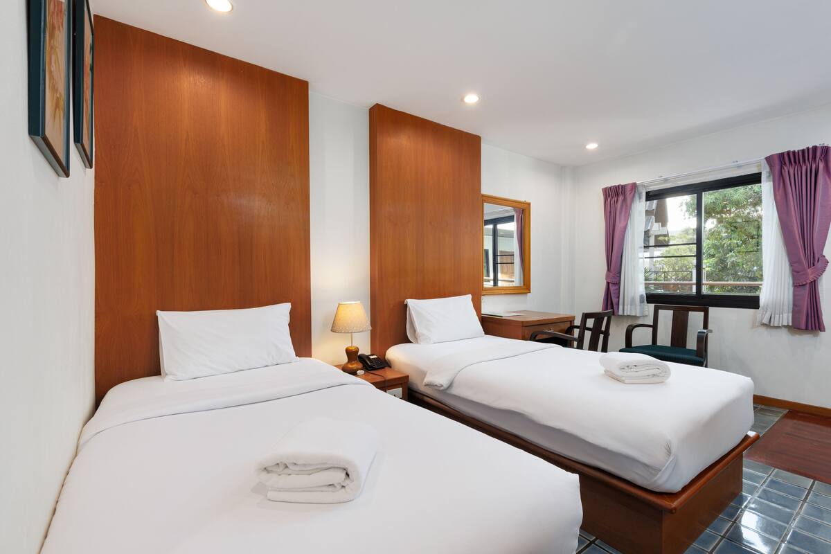 A hotel room with two beds, wooden headboards, a desk with a chair, a window with curtains, and a nightstand with a lamp and phone.