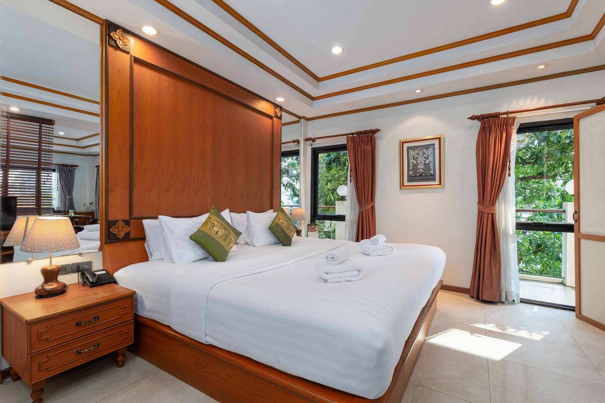 A neatly made bed with white linens and decorative pillows, a wooden nightstand with a lamp and phone, and a large mirror reflecting the room.