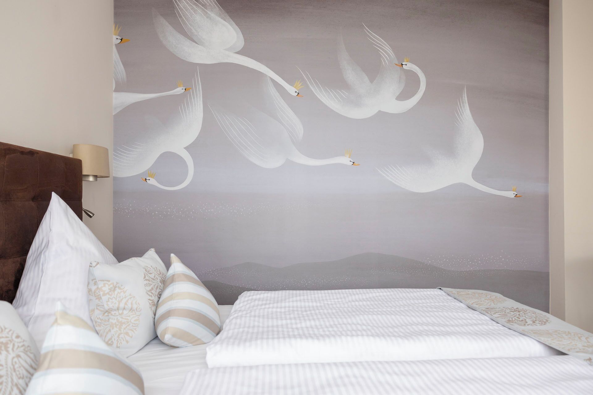 A bedroom with a large wall mural of white swans, a brown headboard, and a bed with white bedding and striped pillows.