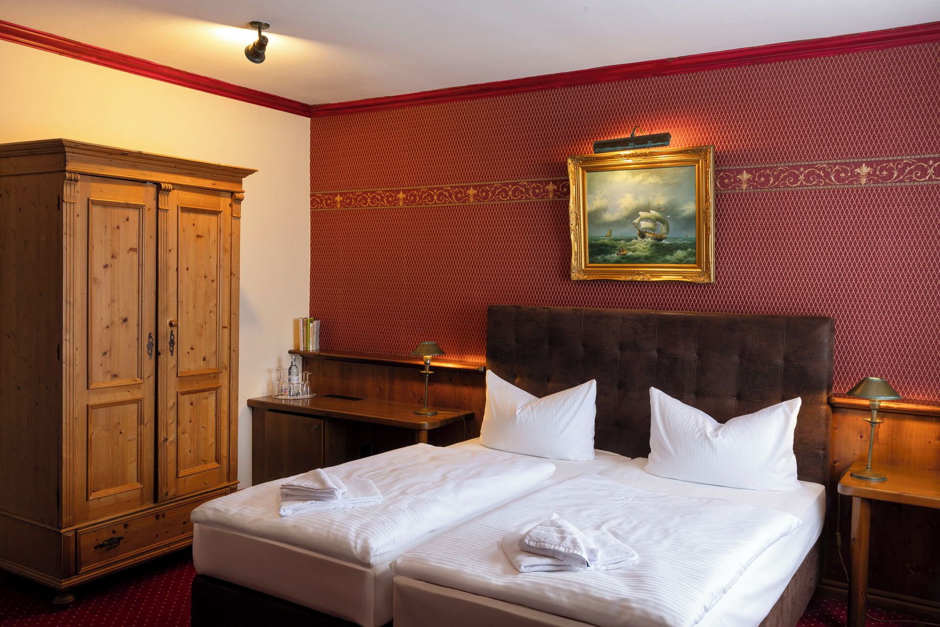 Ein Hotelzimmer mit einem Doppelbett, einem Holzschrank, einem Bild an der Wand und roten Tapete mit Muster.