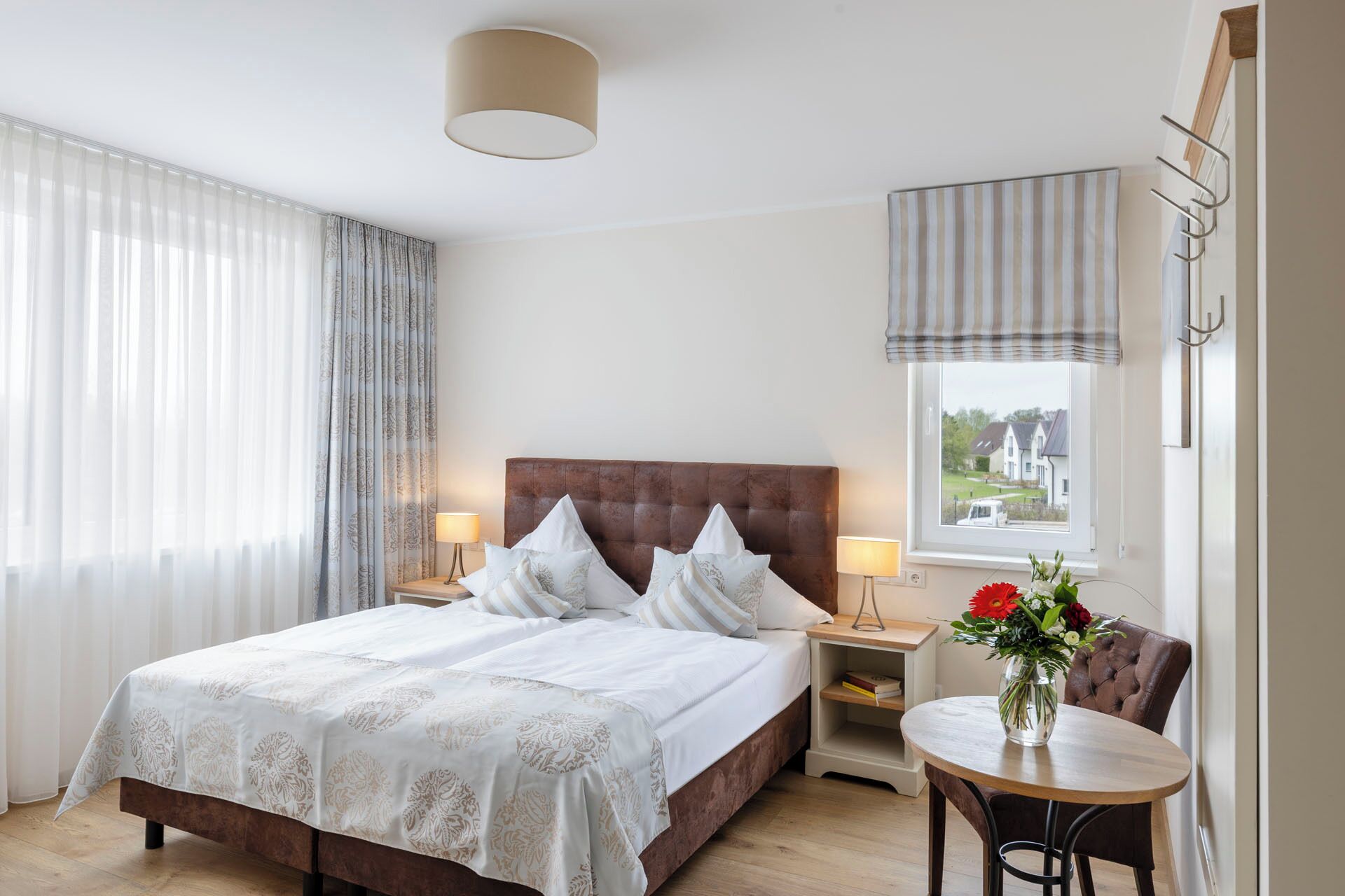 Ett hotellrum med en stor säng, två sänglampor, ett litet bord med en vas med blommor och utsikt över husen genom fönstret.